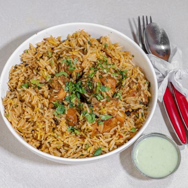Bowl of spiced chicken biryani topped with cilantro, served with creamy green raita and red-handled utensils.
