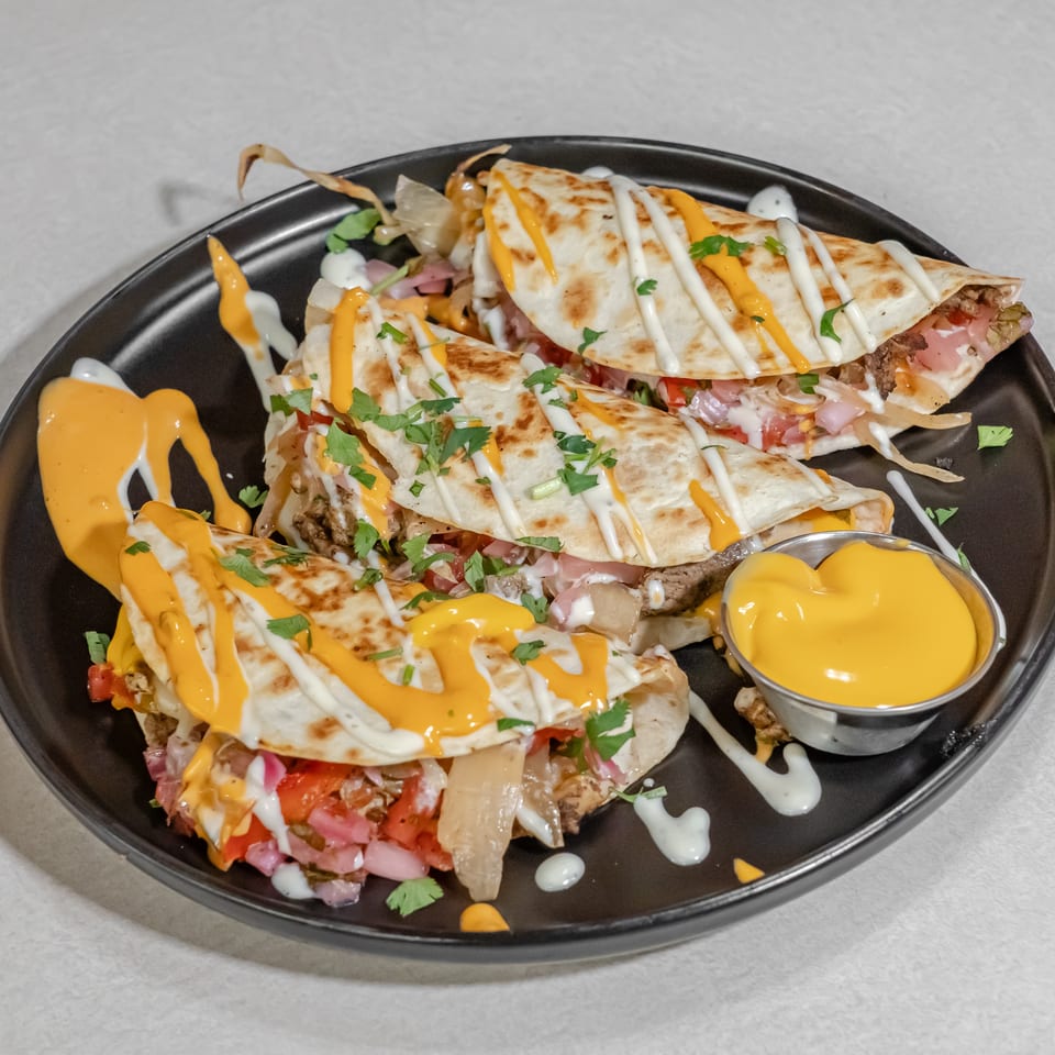 Grilled quesadilla wedges filled with meat and vegetables, drizzled with orange and white sauces, garnished with cilantro, and served with cheese sauce.