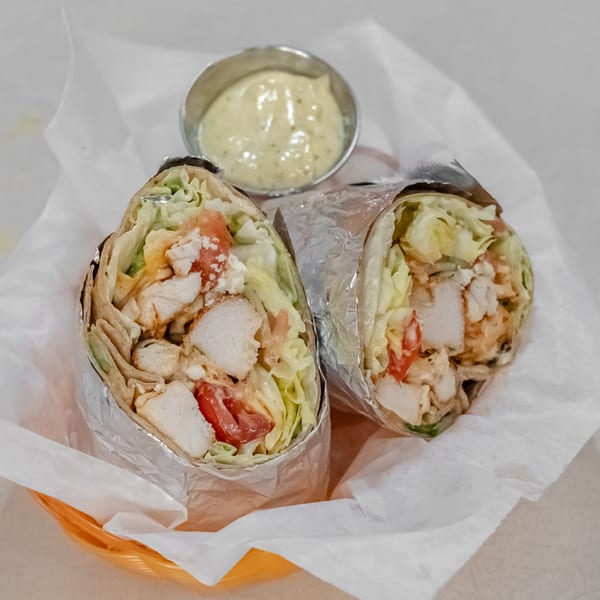 Two grilled chicken wraps filled with lettuce, tomatoes, and sauce, served with a small cup of creamy dipping sauce.