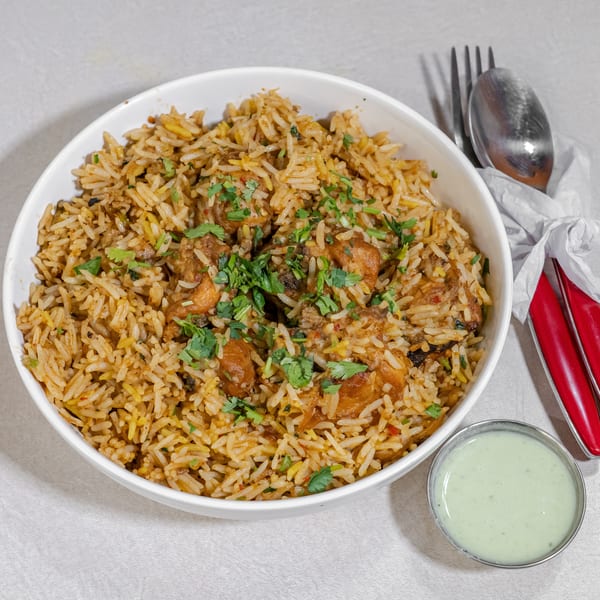 Bowl of spiced rice with roasted chicken pieces and cilantro, served with pale green sauce, spoon, fork, and red-handled utensils.